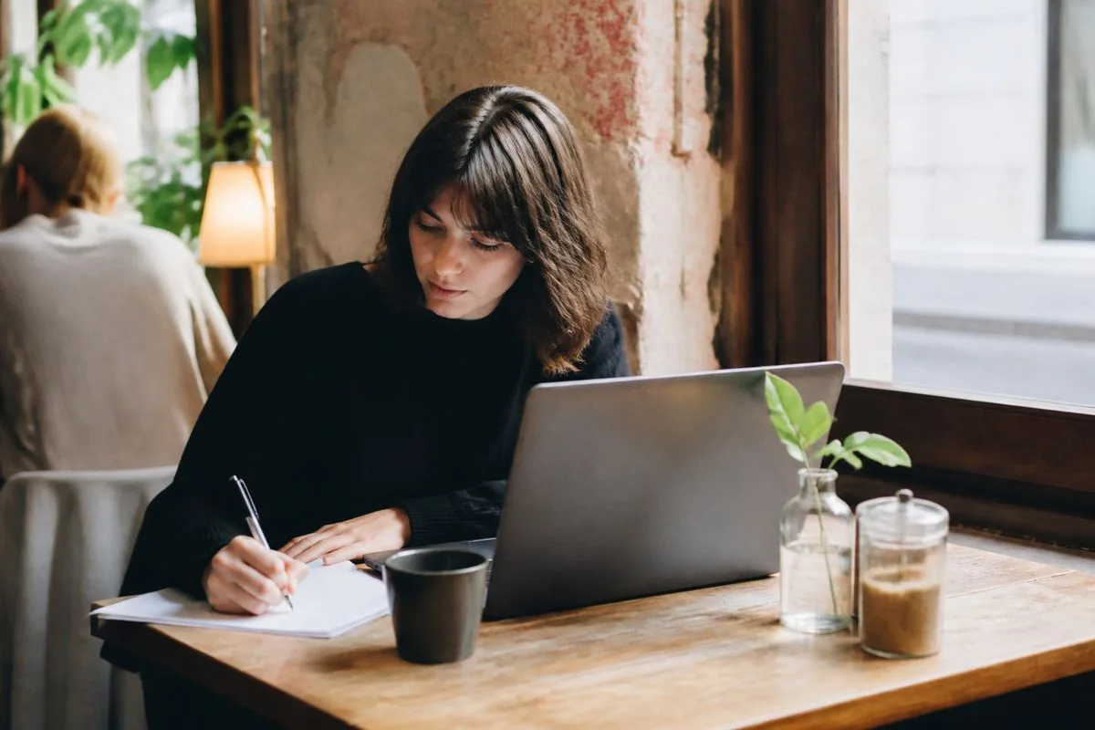 A person using a laptop while taking notes at a café table