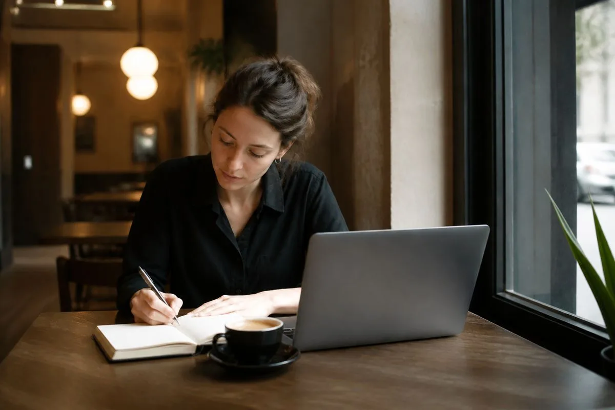 A person comparing notes on a laptop and notebook at a café table