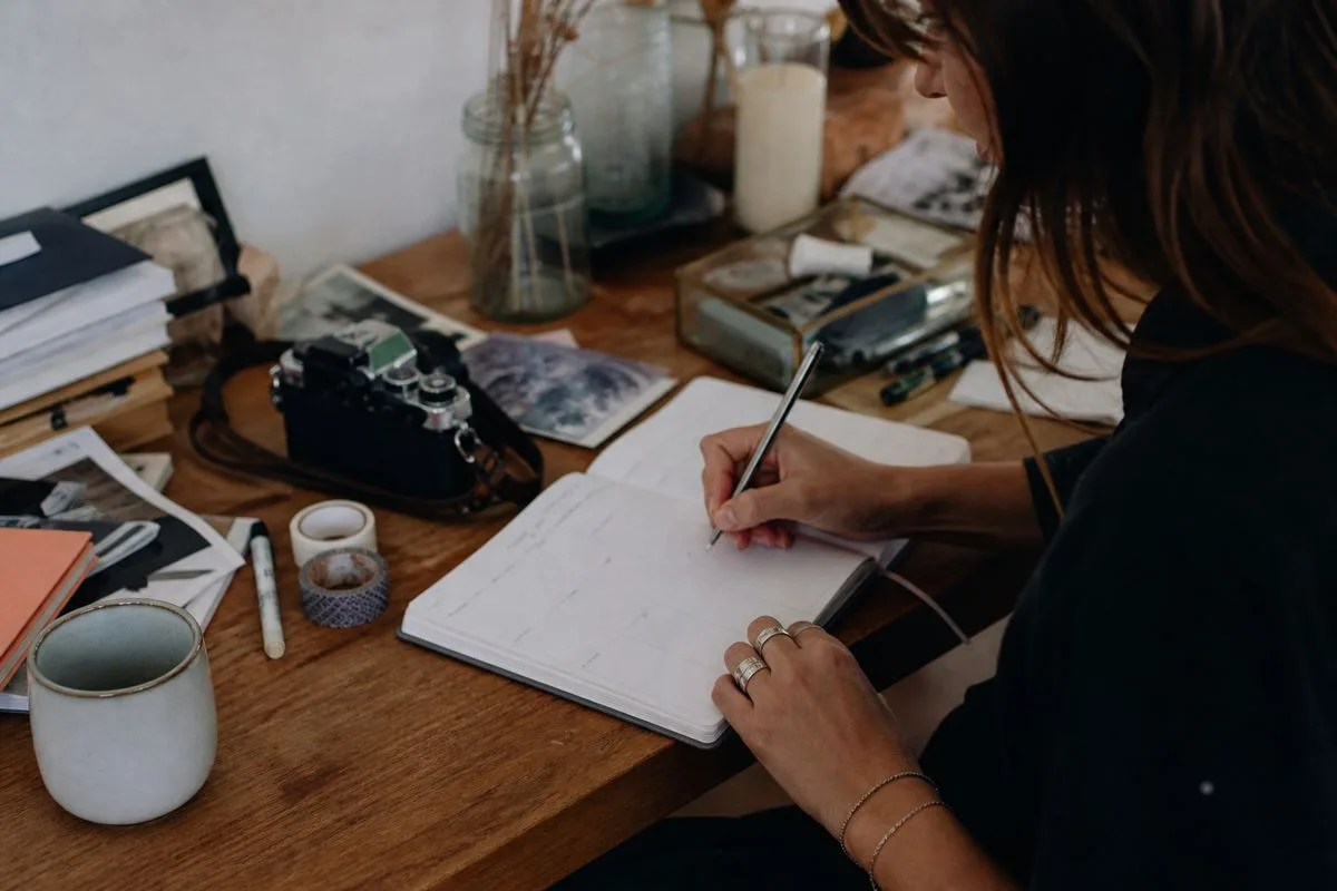 A person writing in a planner at a cluttered desk.